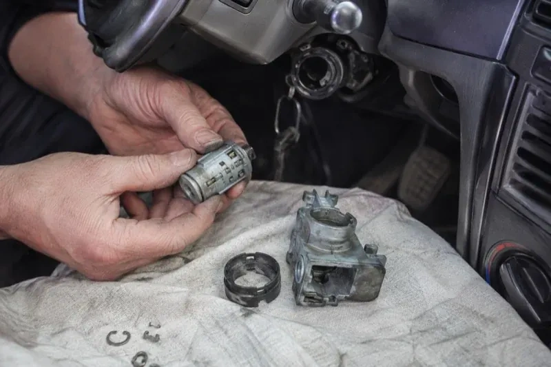 Hands holding a removed ignition cylinder beside a car steering column, with the housing and small parts on a cloth. — Ignition Lock Repair, Richmond Hill